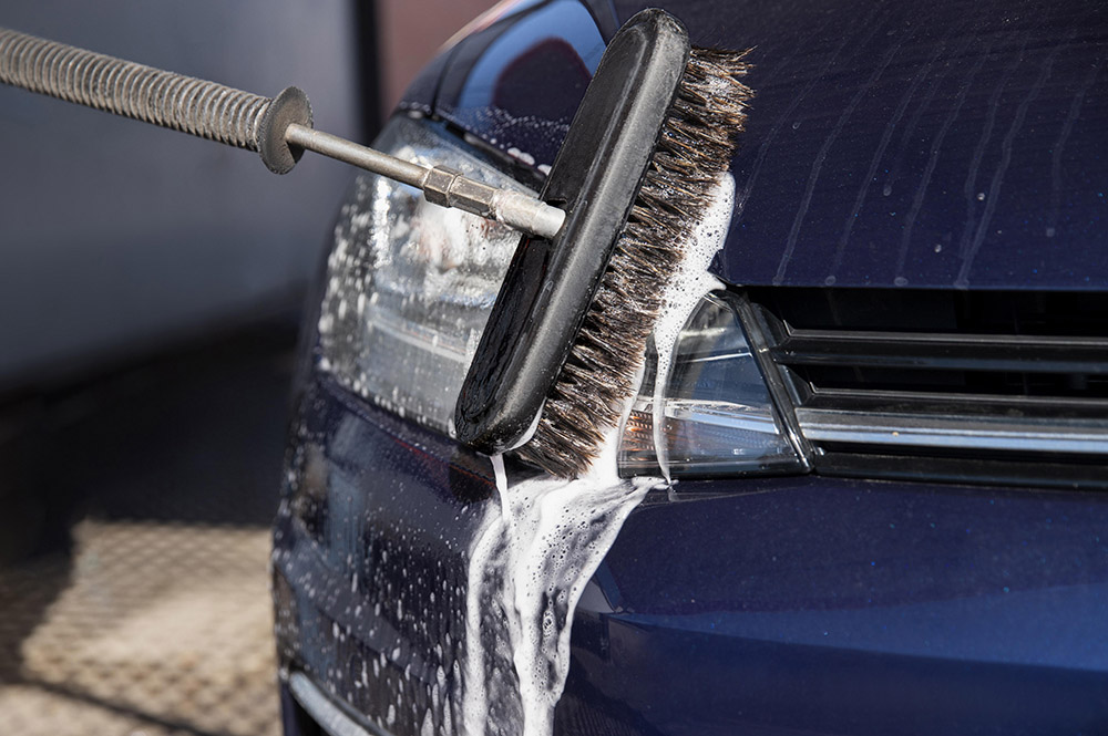 Brosse de lavage couverte de mousse frottant l'avant d'une voiture bleu nuit, la mousse coulant sur le phare
