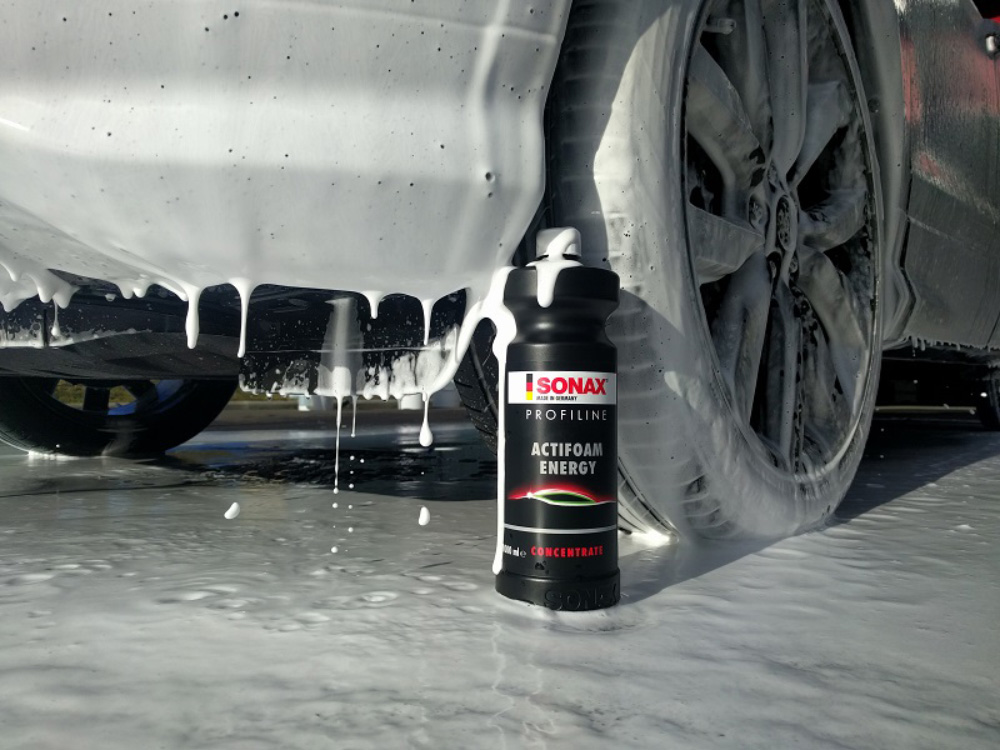 Carrosserie recouverte de mousse de prélavage dégoulinante, bidon SONAX ActiFoam Energy posé au sol devant la roue